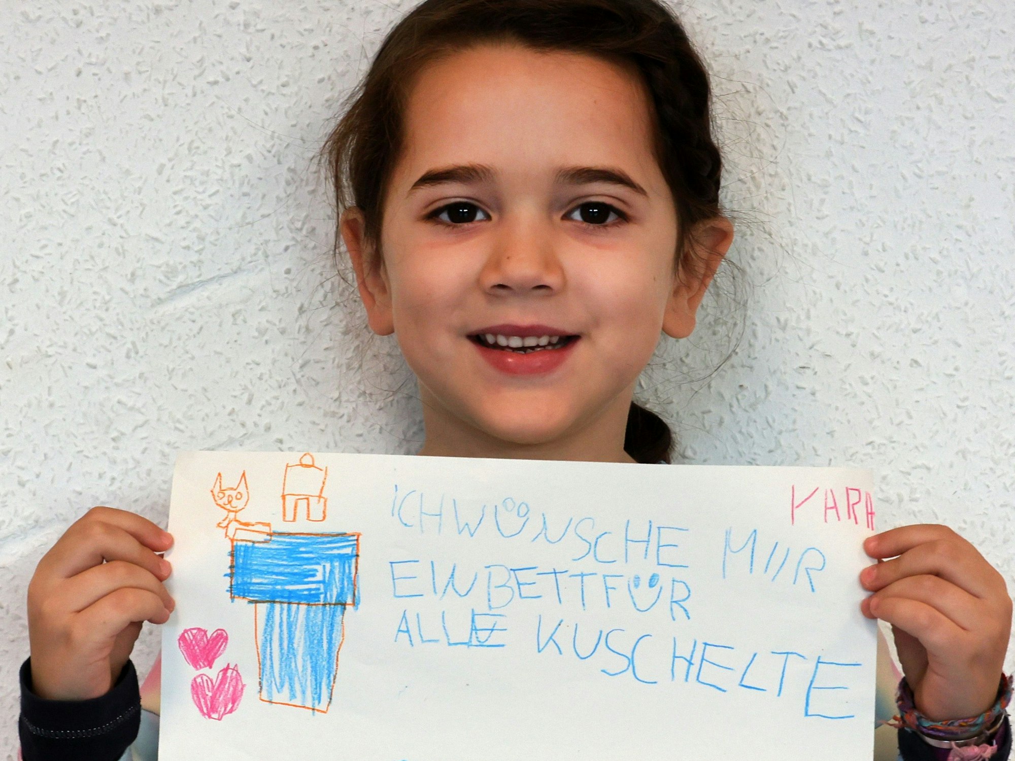 Ein Mädchen hält einen gemalten Wunschzettel in der Hand und blickt in die Kamera.