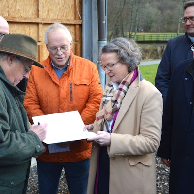 Ditmar Krumpen steht links und unterschreibt den Bescheid. Gegenüber steht die Ministerin für Heimat, Kommunales, Bau und Digitalisierung des Landes Nordrhein-Westfalen Ina Scharrenbach, und Christoph Kammers schaut zu.