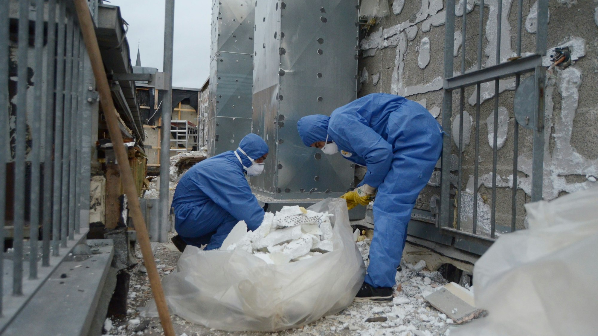 Zwei Männer, die blaue Schutzkleidung und Atemschutzmasken tragen, entfernen Dämmmaterial von der Fassade des City-Forums.