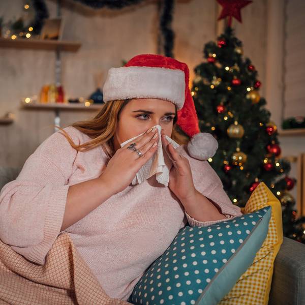 Eine Frau mit Weihnachtsmütze auf dem Kopf liegt auf dem Sofa vor einem Tannenbaum und schnäuzt sich die Nase.