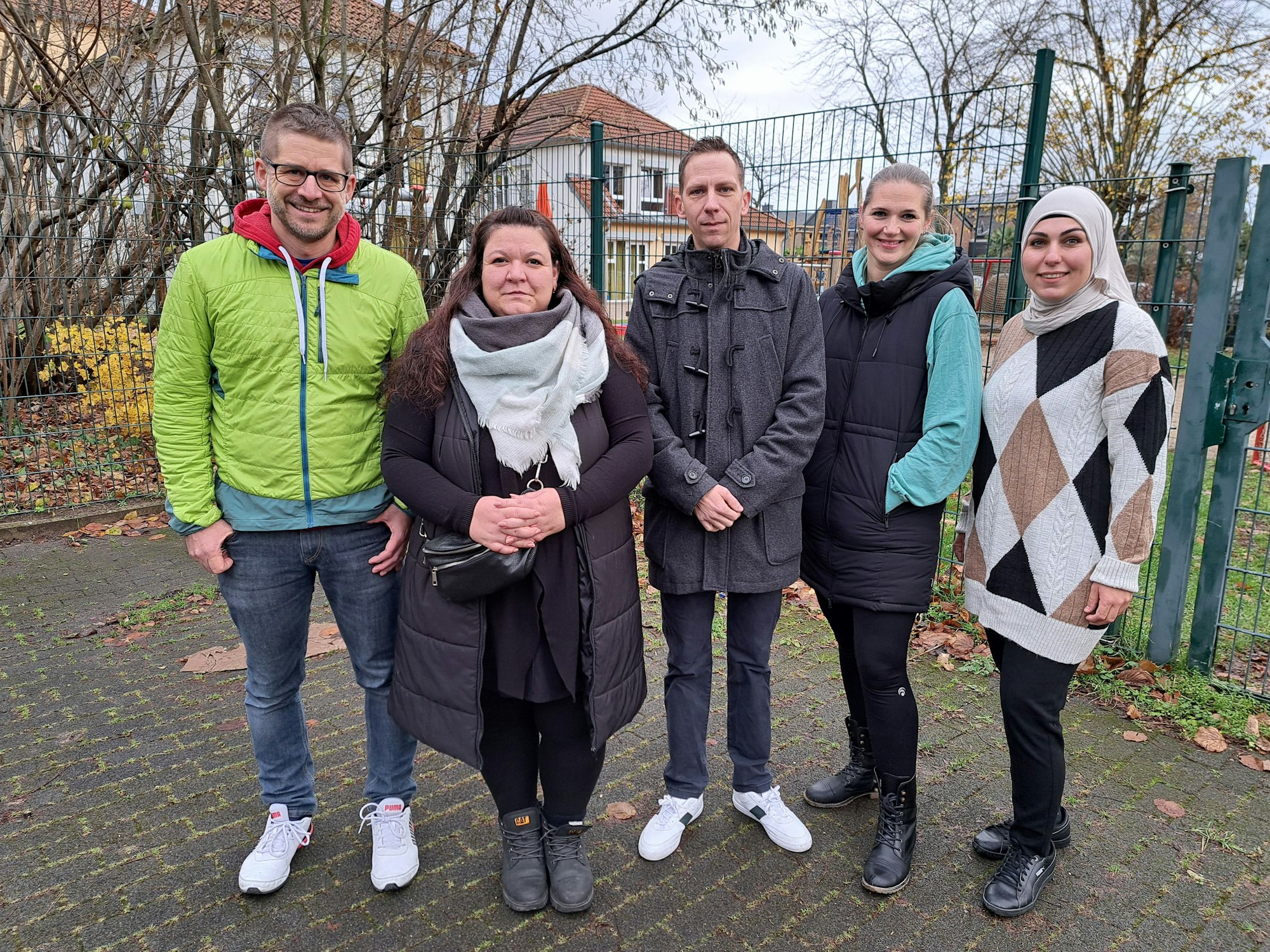 Drei Männer und zwei Frauen stehen vor dem Zaun einer Kita und schauen in die Kamera.
