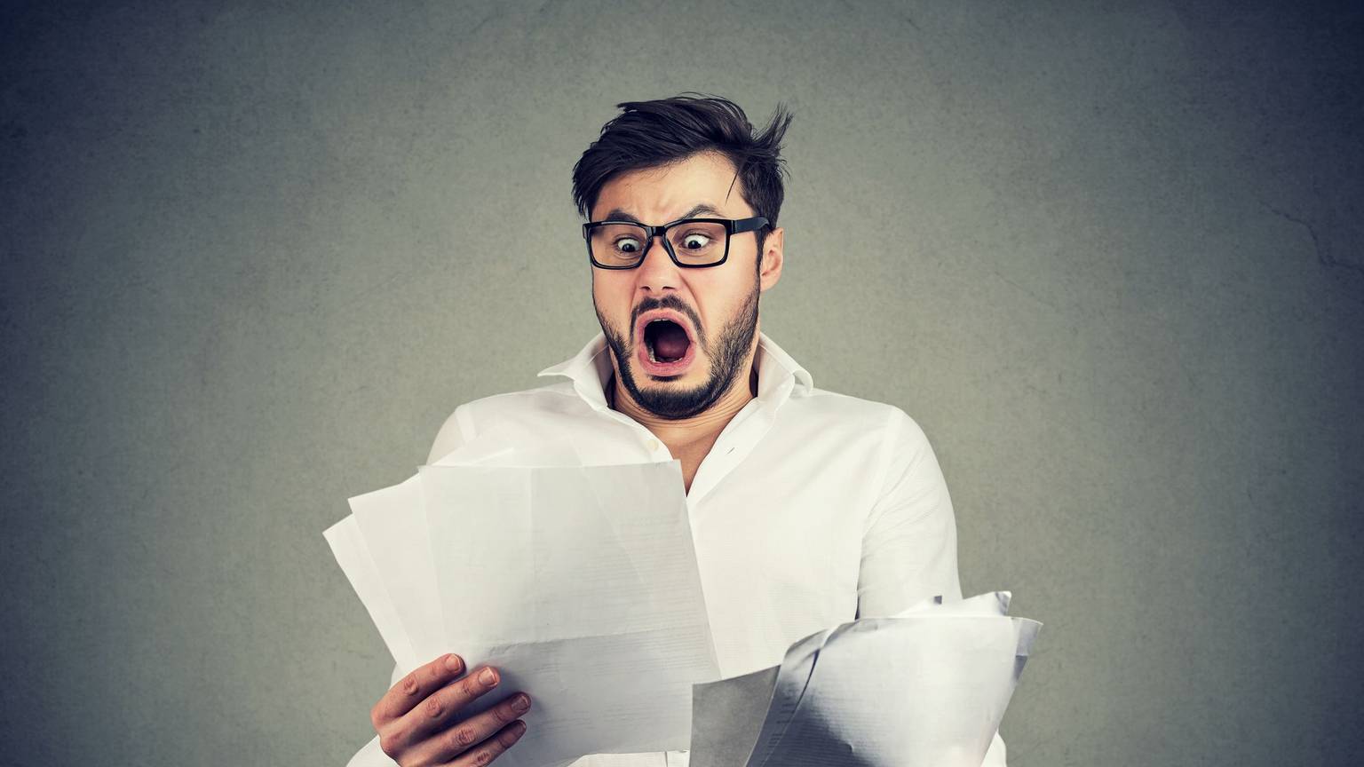 Young shocked guy in glasses looking at papers bank documents with list of bills and fees and feeling stressed on gray background