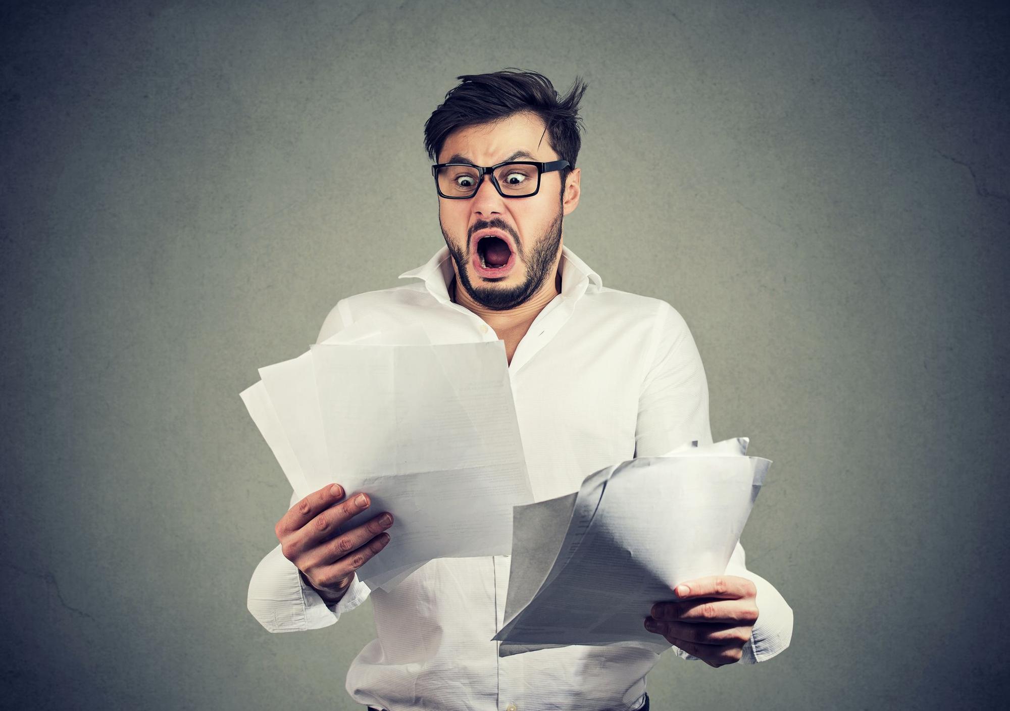 Young shocked guy in glasses looking at papers bank documents with list of bills and fees and feeling stressed on gray background