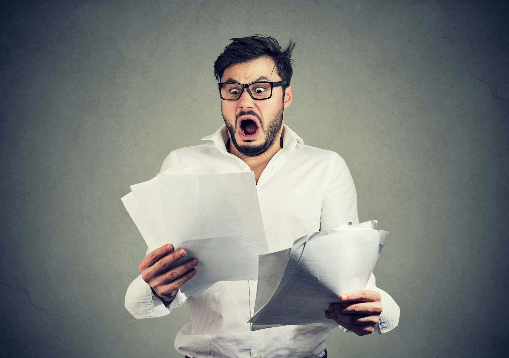 Young shocked guy in glasses looking at papers bank documents with list of bills and fees and feeling stressed on gray background