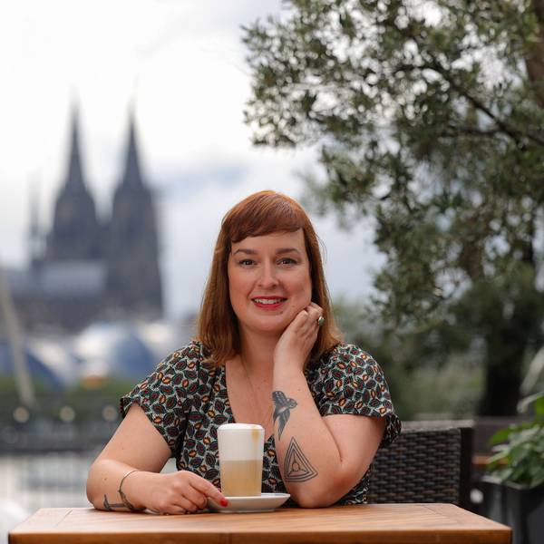 Julia Floß steht mit einem Latte macchiato an einem Stehtisch mit dem Kölner Dom im Hintergrund.