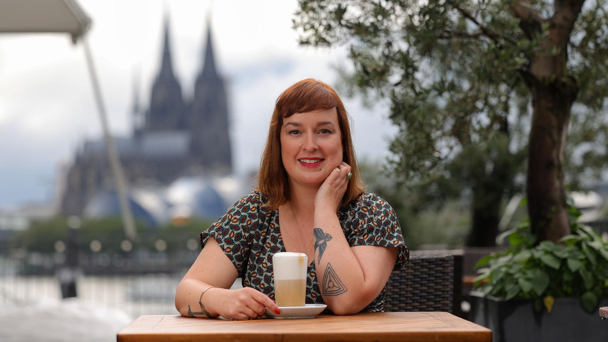 Julia Floß steht mit einem Latte macchiato an einem Stehtisch mit dem Kölner Dom im Hintergrund.
