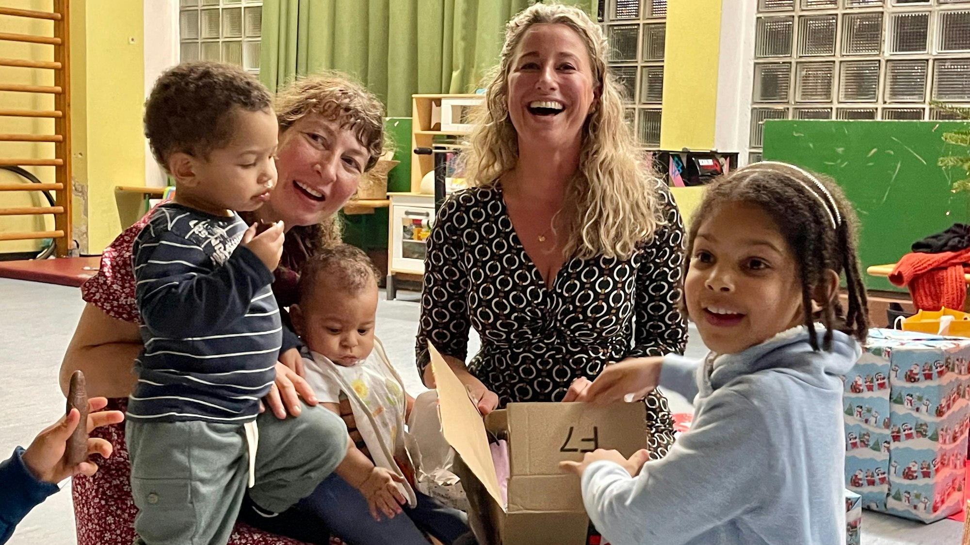 Eine lachende blonde Frau sitzt mit einer Familie mit frei kleinen Kindern auf dem Boden und packt Geschenke aus.