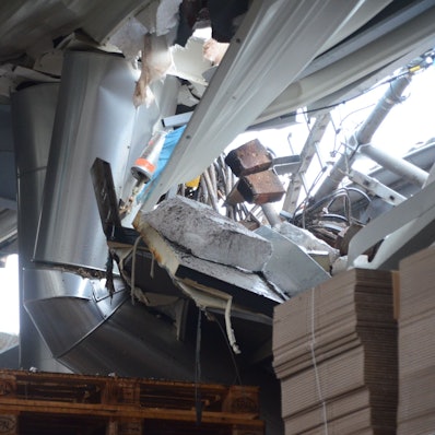 Der Blick aus dem Inneren einer Fabrikhalle durch ein Loch im Dach. Man sieht verbogene Träger und Lüftungsrohre, außerdem einzelne Backsteine und aufgestapelte Kartons.