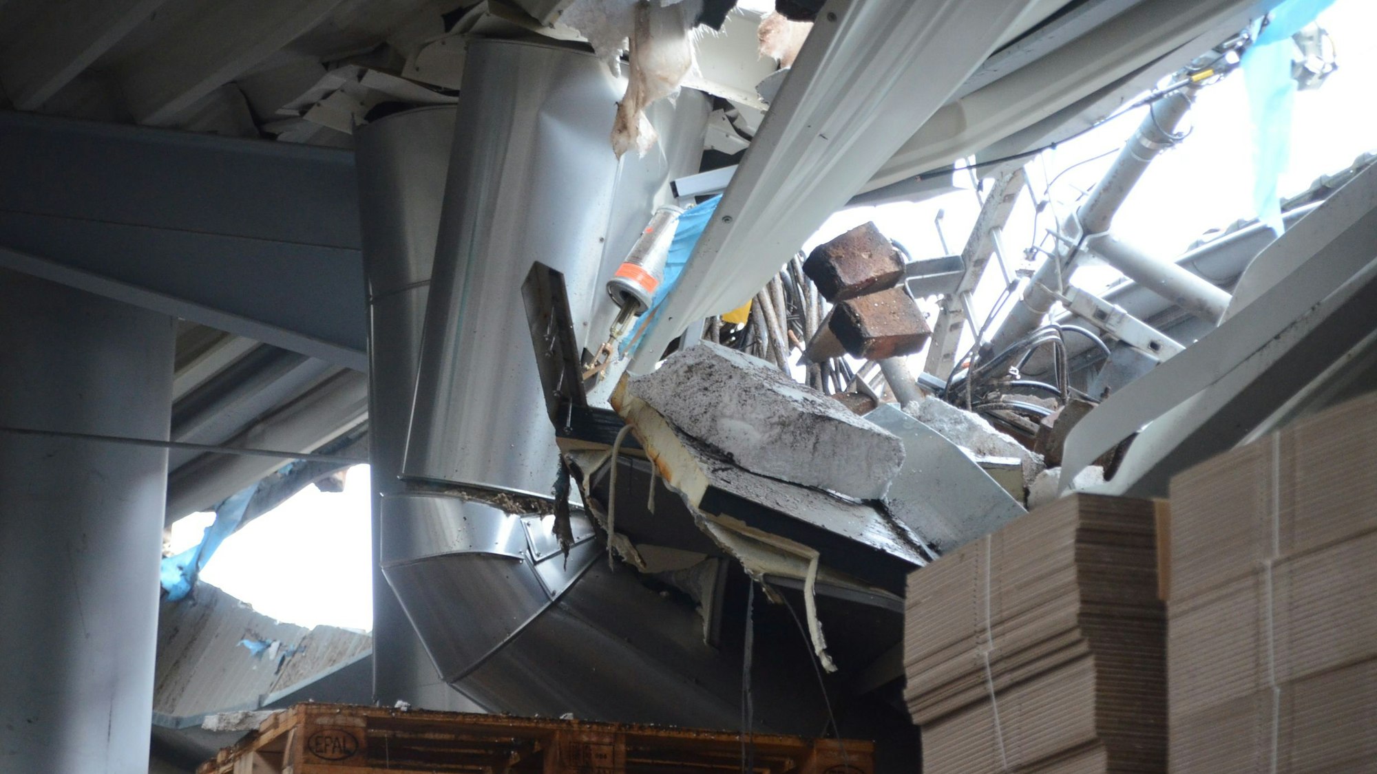 Der Blick aus dem Inneren einer Fabrikhalle durch ein Loch im Dach. Man sieht verbogene Träger und Lüftungsrohre, außerdem einzelne Backsteine und aufgestapelte Kartons.