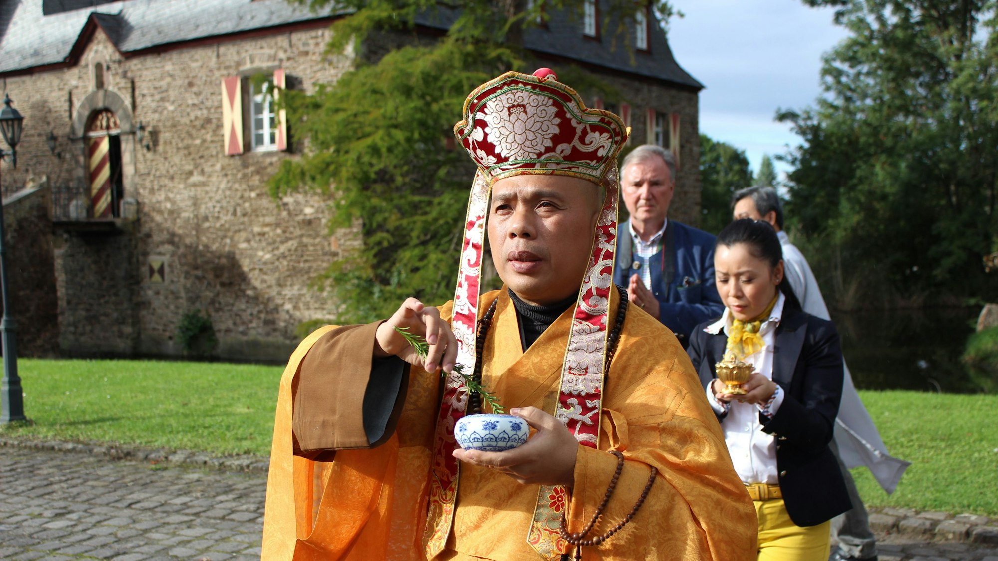 Ein festlich gekleideter Mönch in orangefarbenem Gewand schreitet voran, dahinter der Freiherr und seine Lebensgefährtin, im Hintergrund das Herrenhaus.
