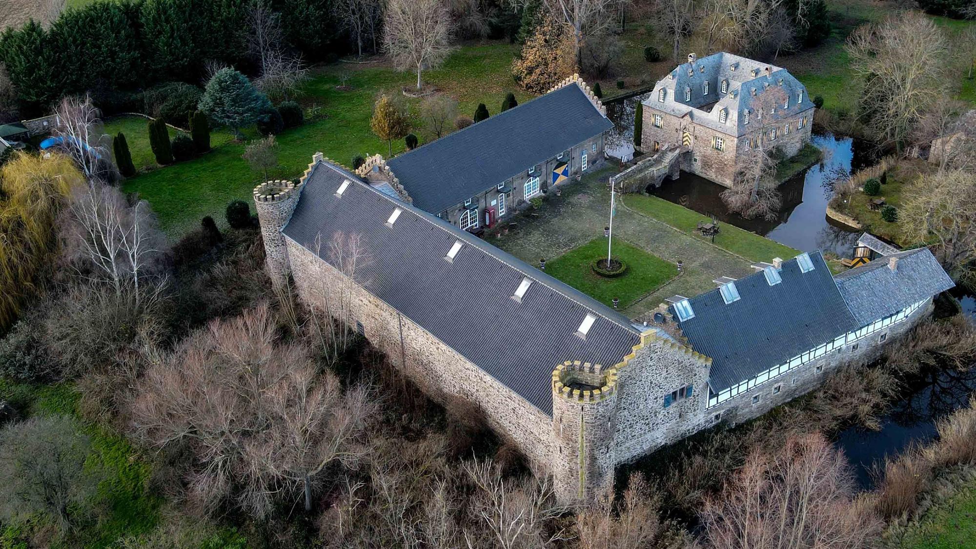 Das Foto zeigt das Burg-Ensemble aus der Luft. Im Vergleich zu den einen Hof umgebenden Wirtschaftsflügeln mit den beiden runden Türmen der Vorburg ist das über eine Zugbrücke zu erreichende Herrenhaus relativ klein. Umgeben ist die Anlage von Wassergräben und einem Park.