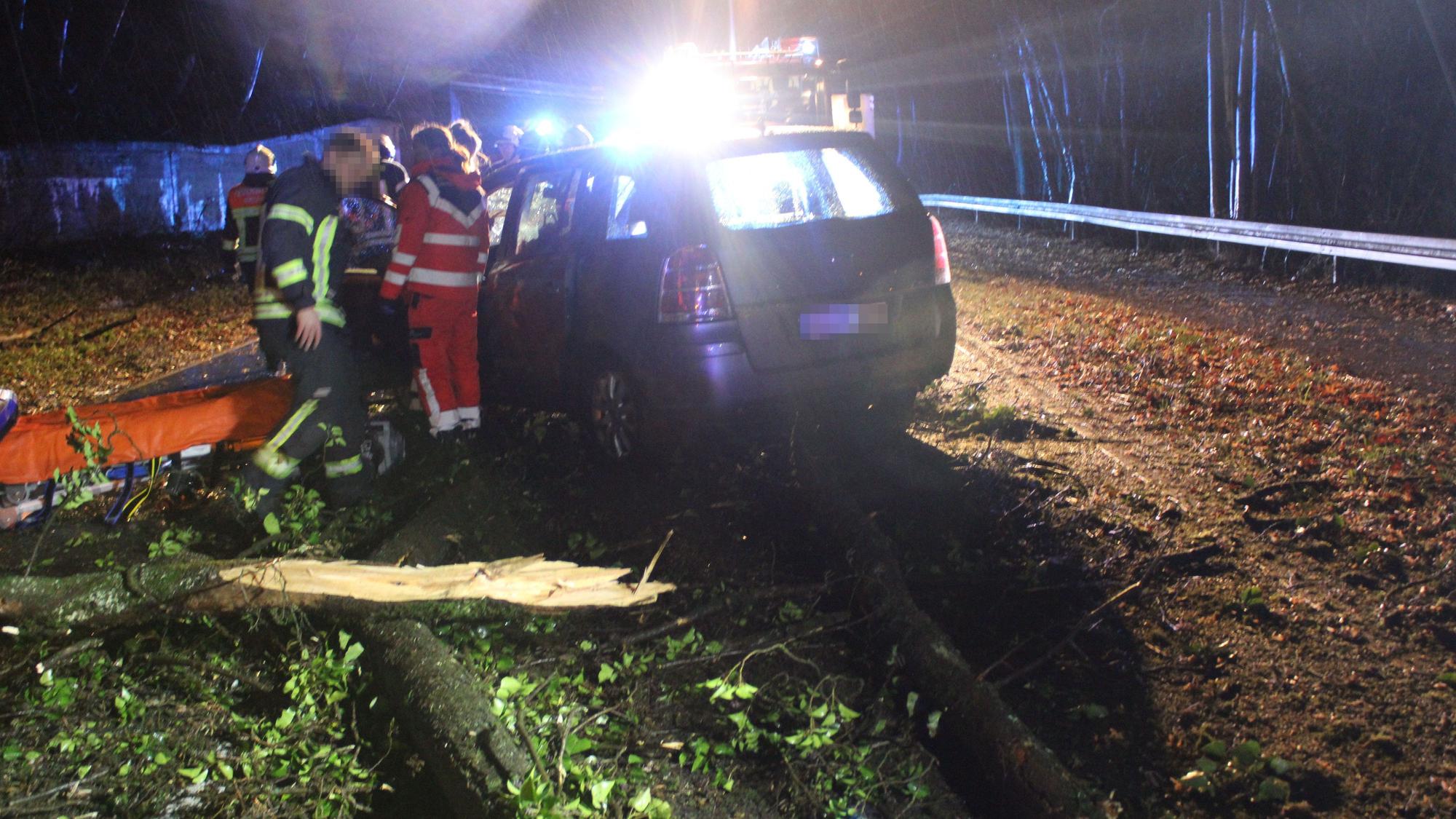 Ein Unfallauto steht vor einem umgestürzten Baum, im Hintergrund ist Blaulicht zu sehen.
