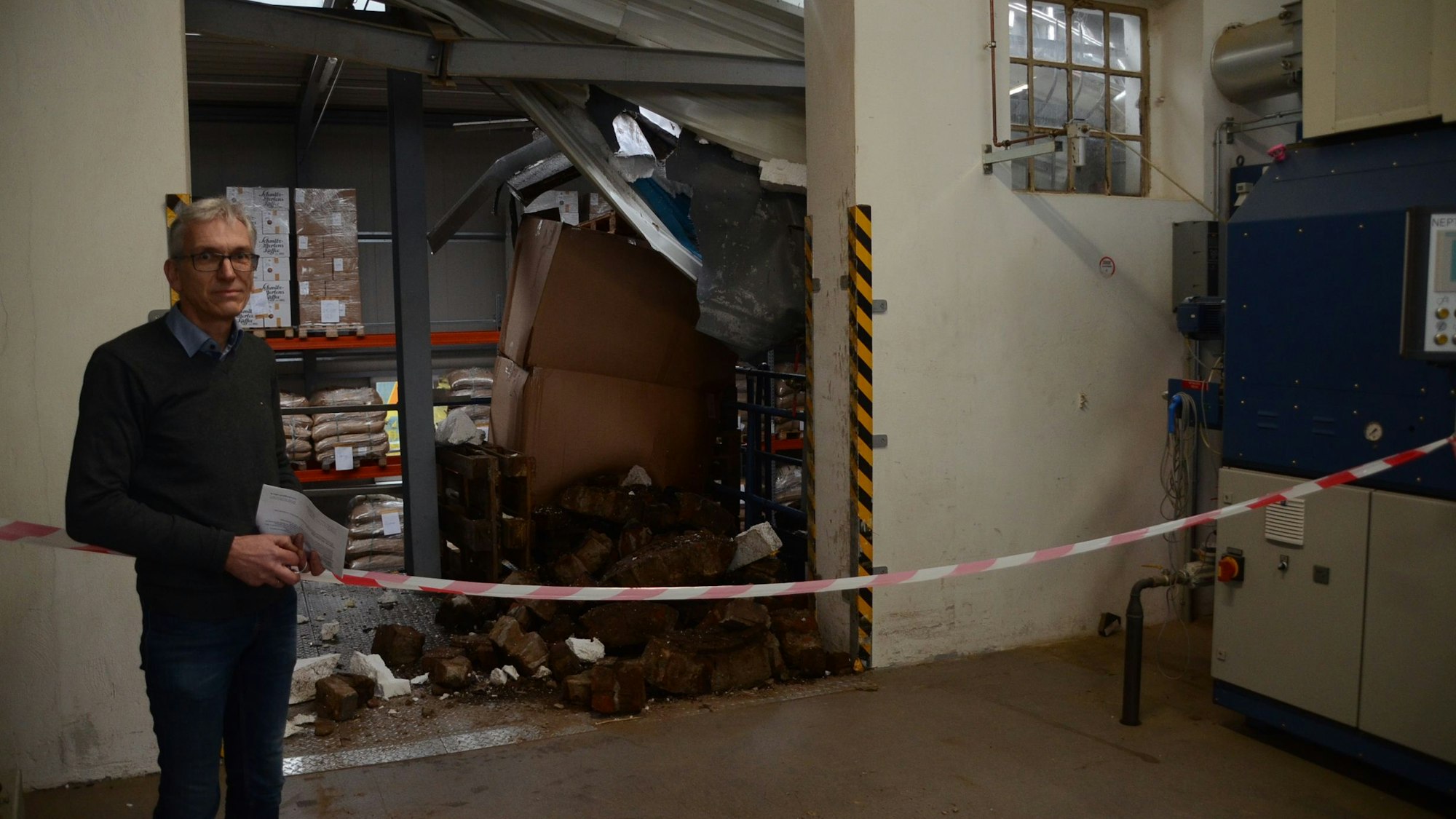 Ein Mann mit Brille und grauen Hallen steht vor einem Haufen an Backsteinen und verbogenem Metall.
