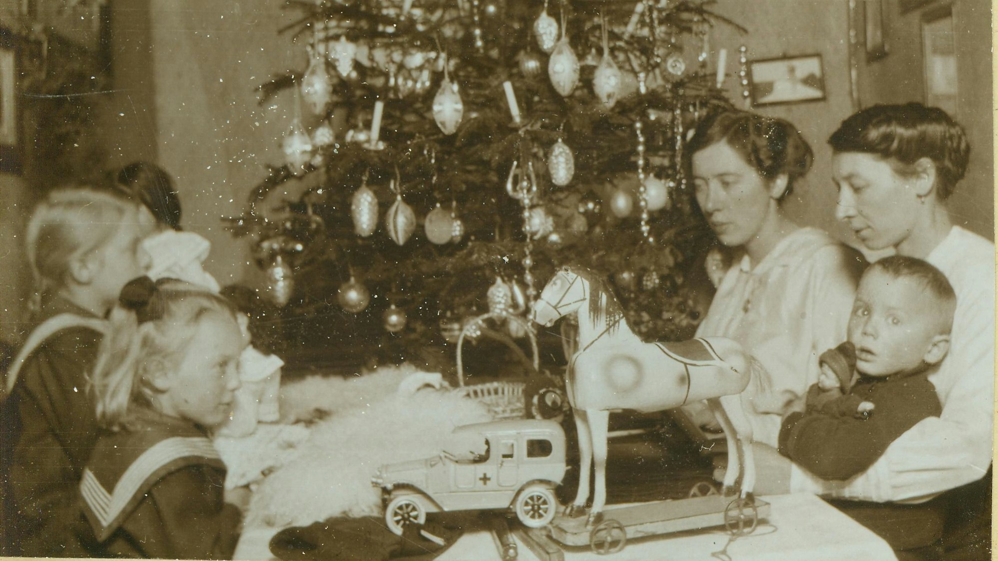 Weihnachtsfest vor mehr als 100 Jahren: Eine Familie sitzt vor einem geschmückten Weihnachtsbaum. Auf dem Tisch stehen Geschenke und Spielzeug für die Kinder.