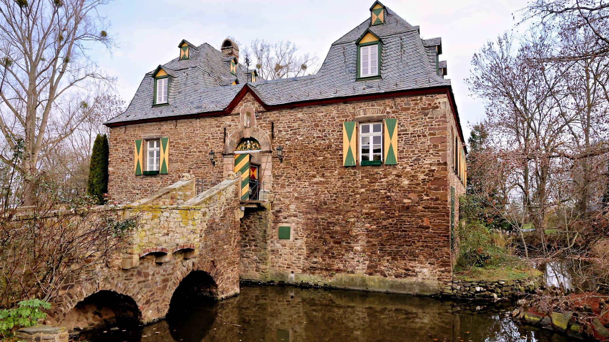 Blick über den Wassergraben hinweg auf das Herrenhaus, links die Brücke und die herabgelassene Zugbrücke.
