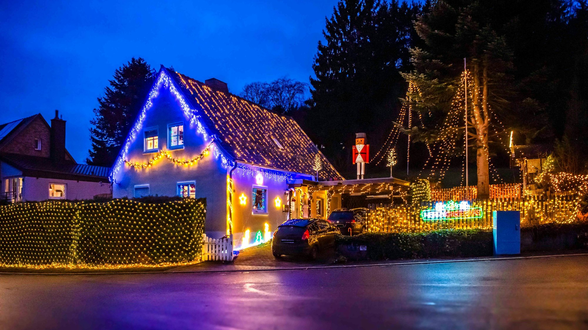 Ein mit Lichterketten behangenes Haus steht an einer Straße. Auf der Garage steht eine große Nussknacker-Figur. Auch Garten, Hecke und Zaun sind mit Lichterketten geschmückt.