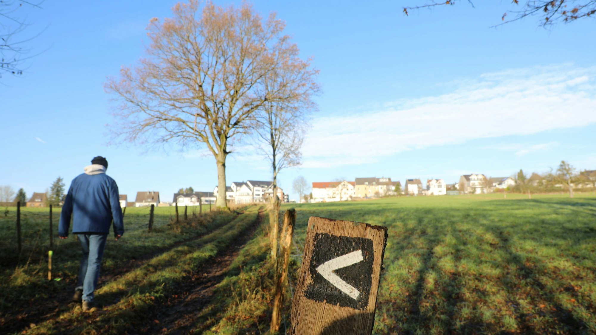 Ein Wanderer geht über einen Wiesenweg, neben dem ein Pfosten mit einer Wegmarkierung steht.