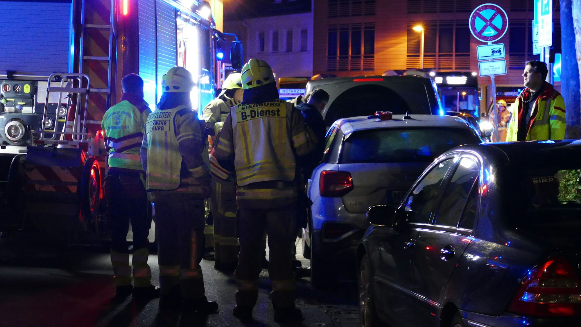Rettungskräfte und ein Feuerwehrfahrzeug stehen in einer Straße mit parkenden Fahrzeugen.
