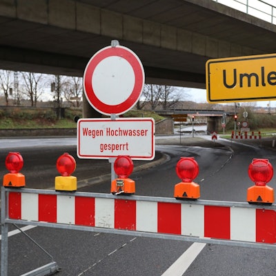 Eine rot-weiße Warnbake mit dem Hinweis auf das Hochwasser steht auf einer Straße.