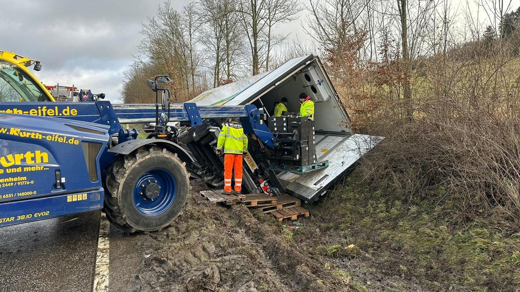 Der Lkw liegt im Straßengraben, Mitarbeiter des Bergungsunternehmens stapeln die Kisten vor dem Wagen.