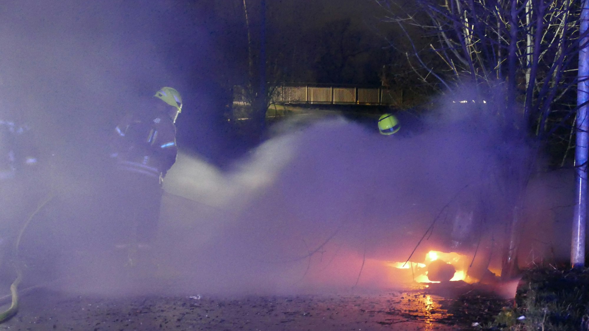 Feuerwehrleute stehen in dichtem Qualm und löschen, heller Flammenschein ist zu sehen.