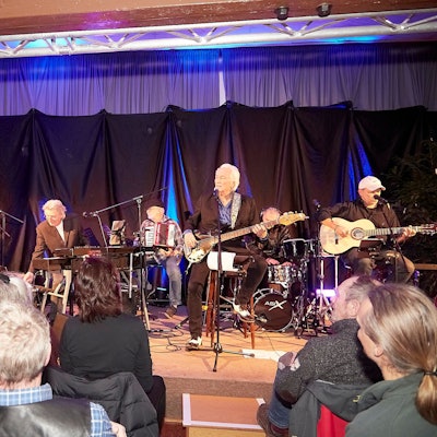 Hannes Schöner sitzt auf einem Hocker, spielt Bass und singt. Im Hintergrund sind weitere Musiker zu sehen.
