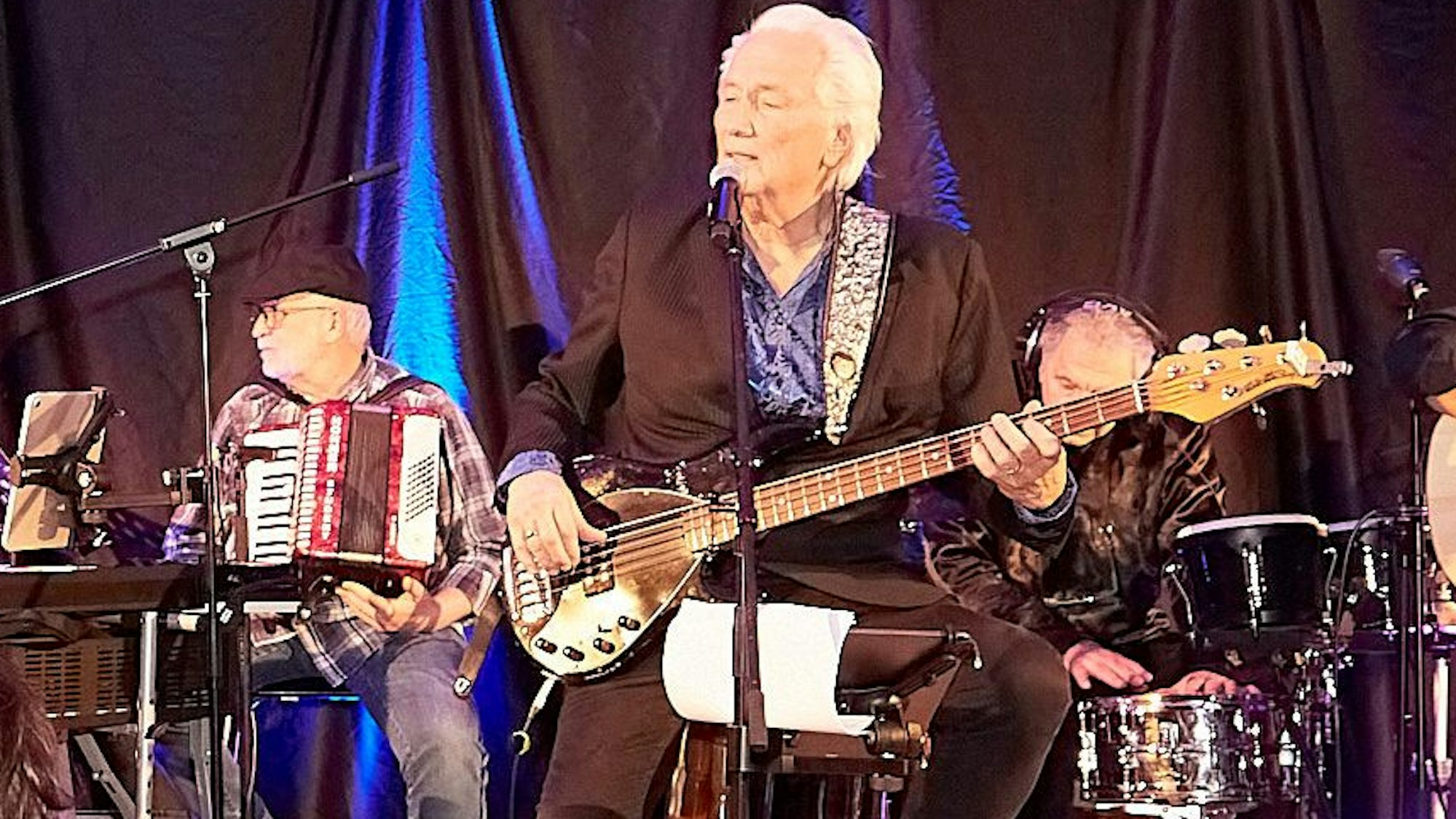 Hannes Schöner sitzt auf einem Hocker, spielt Bass und singt. Im Hintergrund sind weitere Musiker zu sehen.