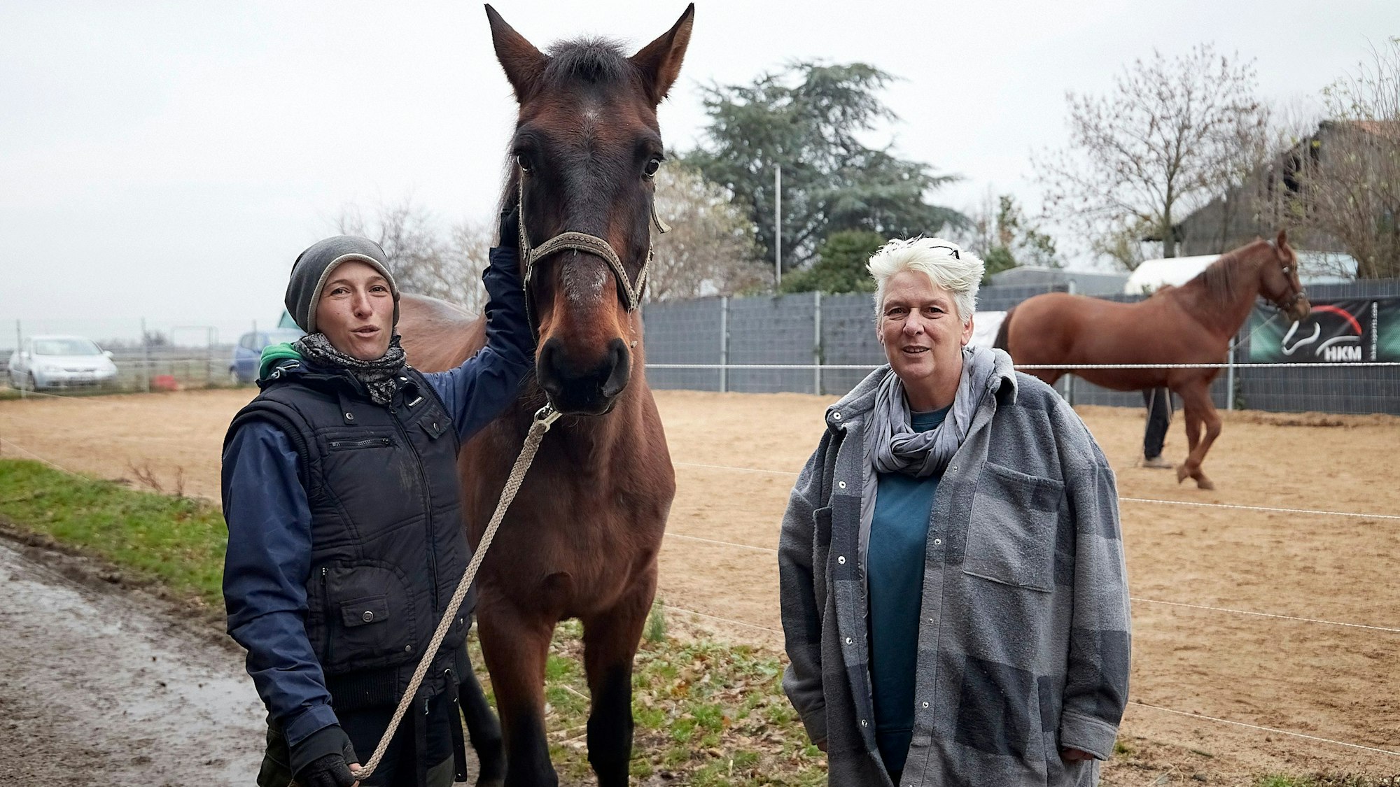 Nina Aschhoff und Bettina Kossmann stehen an einem Außenreitplatz. Nina Aschhoff hält ein Pferd am Halfter.