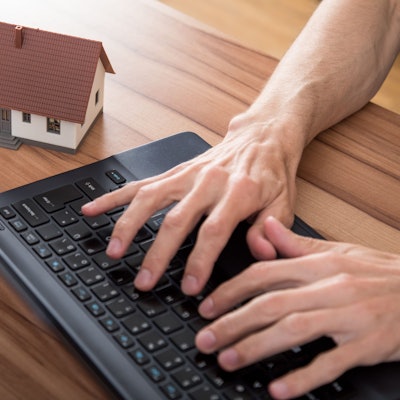 Hände tippen auf eine Tastatur, daneben steht ein Miniaturmodell eines Hauses auf dem Tisch.