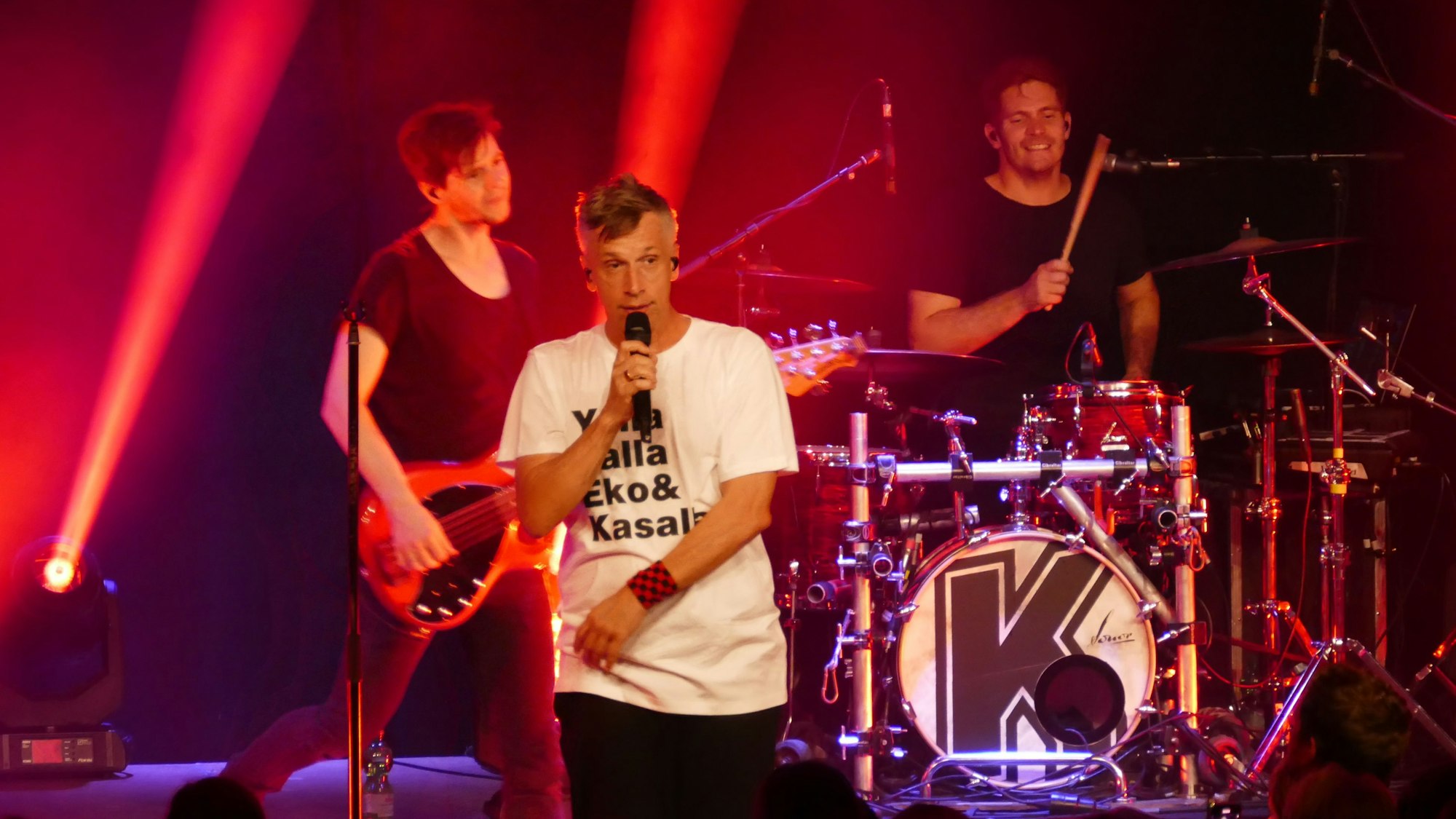 Die Band Kasalla ist bei einem Auftritt auf der rot beleuchteten Bühne zu sehen. Sänger Basti Campmann trägt ein weißes T-Shirt.