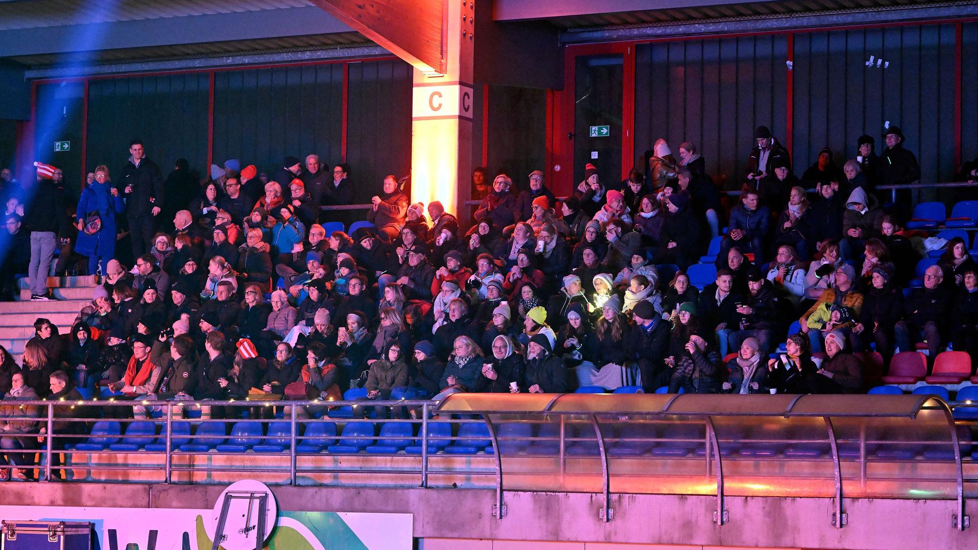 Menschen sitzen auf der illuminierten Tribüne des Bergisch Gladbacher Stadions und singen.