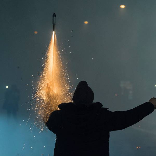 Eine Person hält auf einer Straße in der einen Hand eine zündende Rakete, in der anderen einen Böller, im Hintergrund Rauchschwaden von Feuerwerk.
