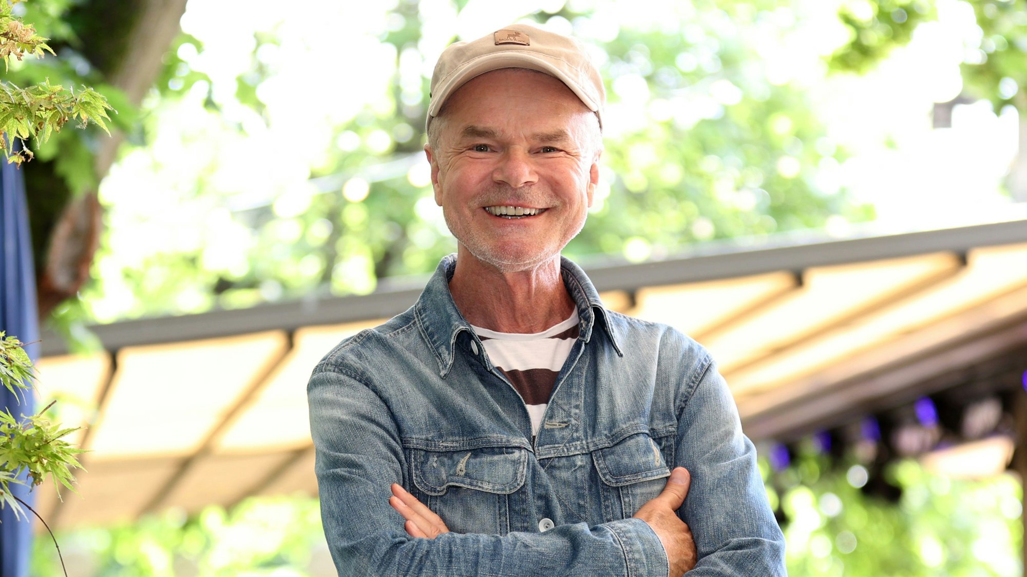 Autor und Moderator Jürgen Domian ist im Halbporträt in sommerlicher Umgebung zu sehen. Er hat die Arme vor der Brust verschränkt, trägt eine Jeansjacke überm T-Shirt und eine beige Baseball Cap.