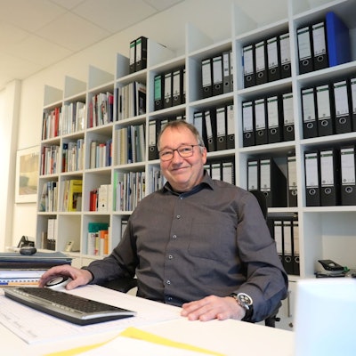 Ein Mann sitzt am Schreibtisch eines Büros, vor sich eine Tastatur. Im Hintergrund steht ein Regal voller Aktenordner.