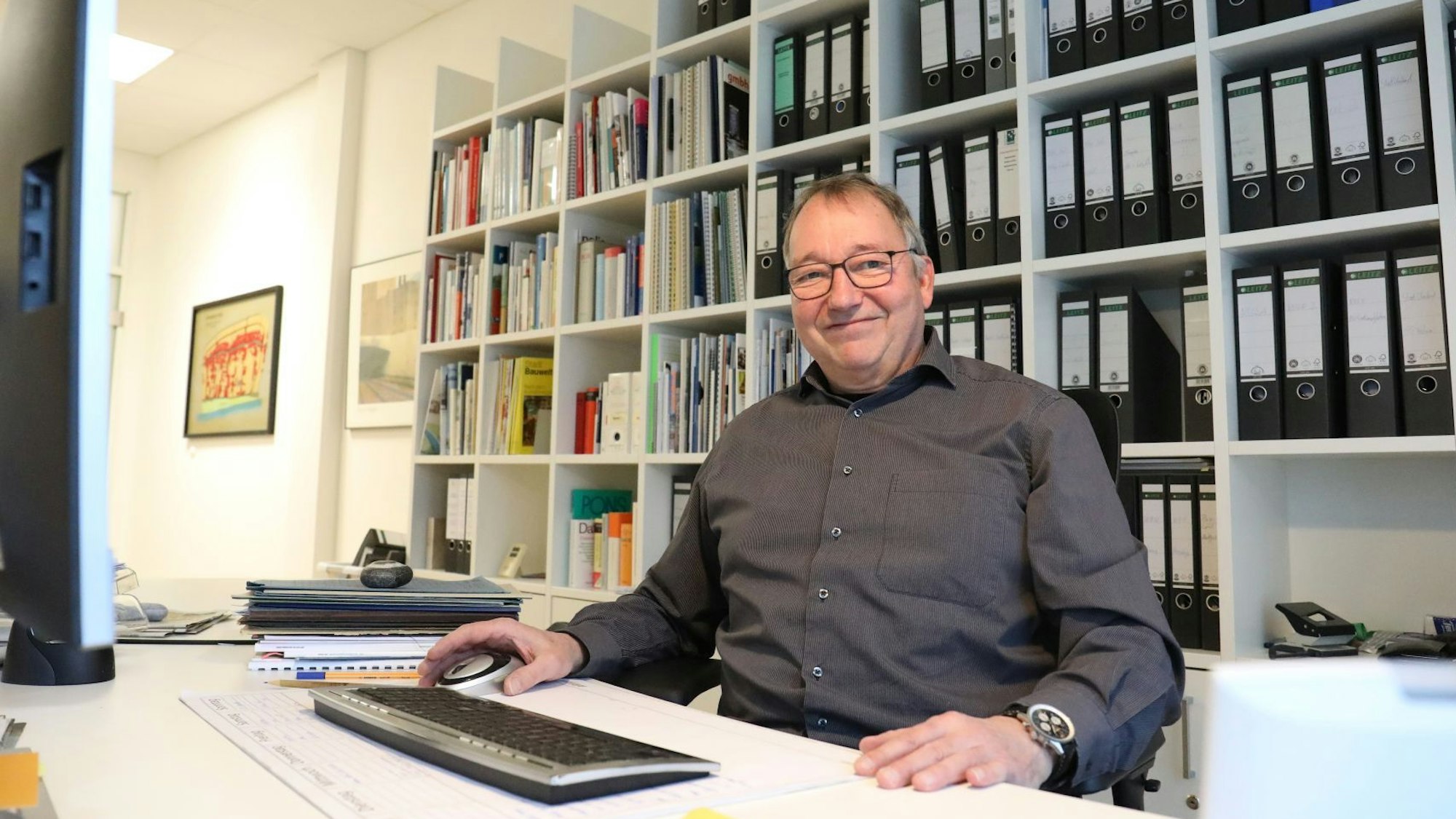 Ein Mann sitzt am Schreibtisch eines Büros, vor sich eine Tastatur. Im Hintergrund steht ein Regal voller Aktenordner.