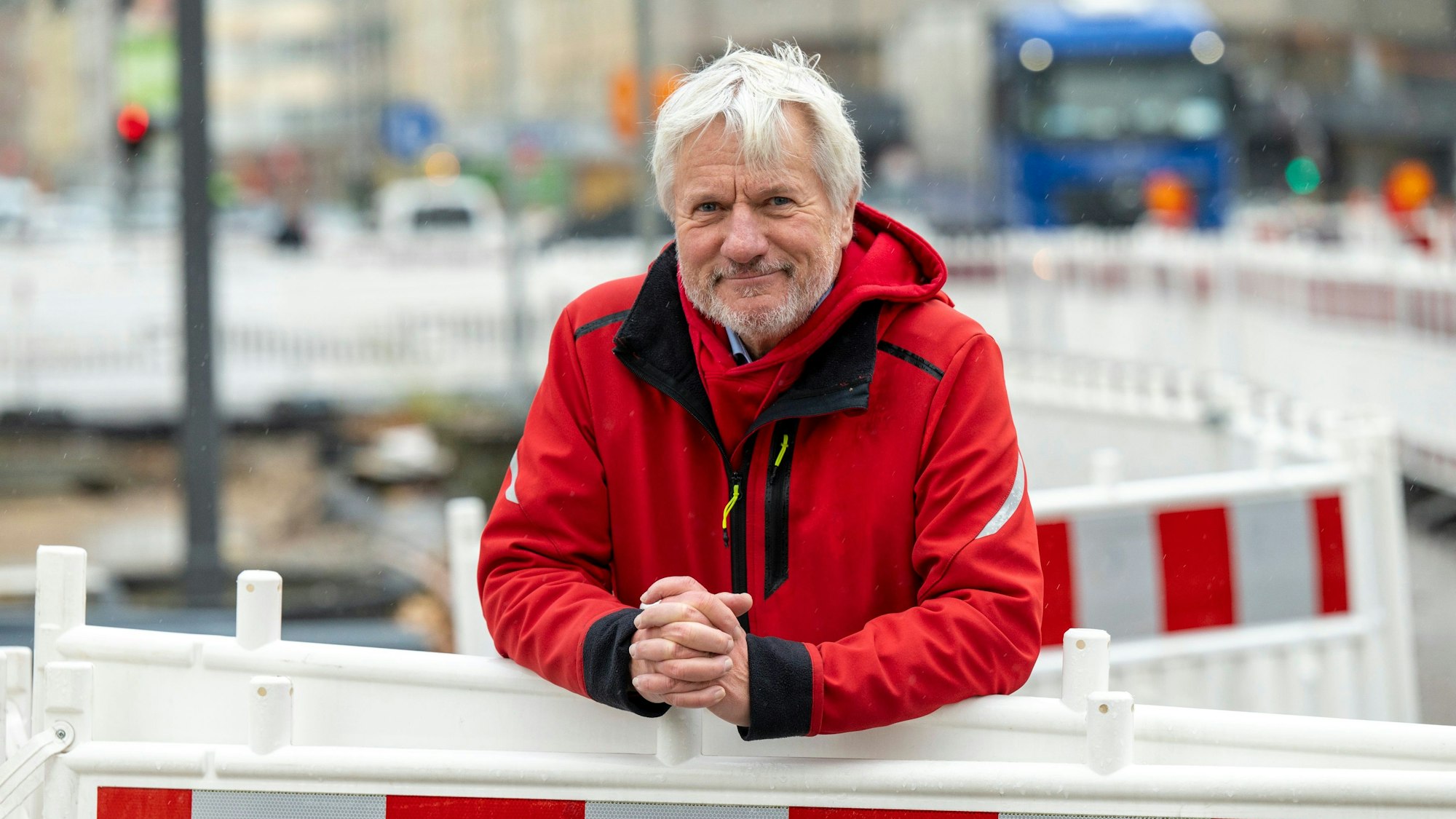 Jürgen Becker steht hinter einer halbhohen Bauabsperrung und schaut freundlich in die Kamera. Er trägt eine knallrote Winterjacke.