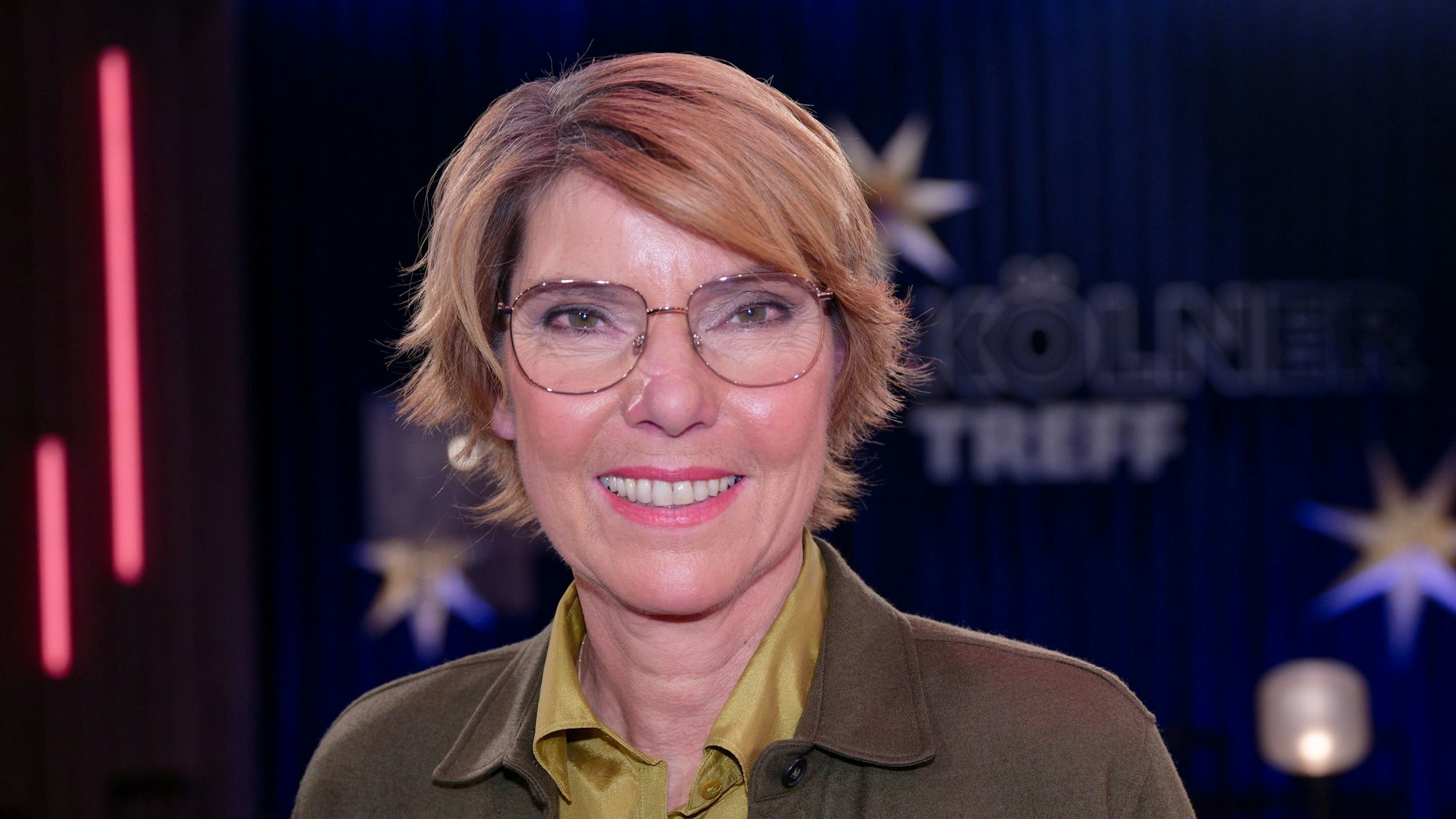 Die Moderatorin Bettina Böttinger lacht in die Kamera. Im Hintergrund ist die Kulisse ihrer früheren Show Kölner Treff zu sehen.