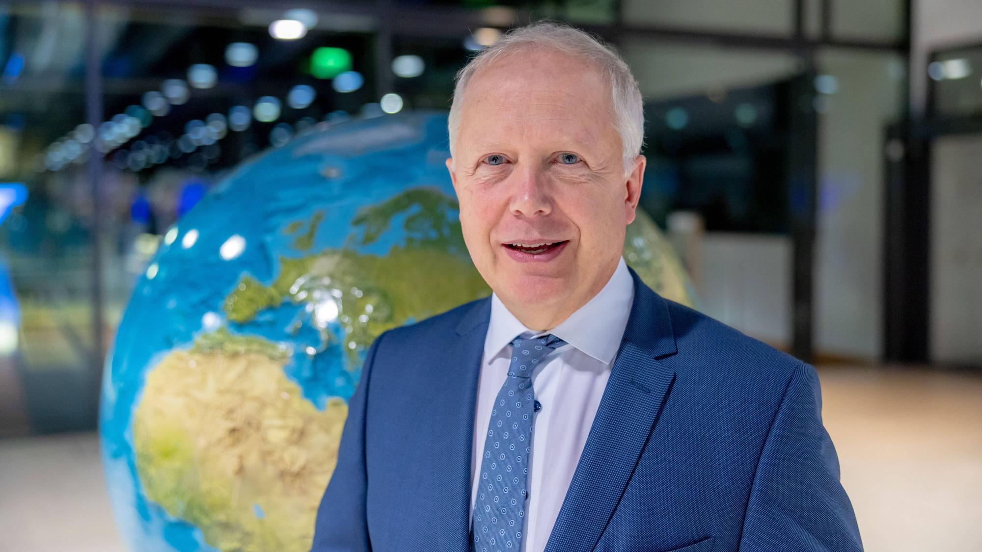 WDR-Intendant Tom Buhrow steht vor einer Weltkugel. Er trägt einen blauen Anzug und lächelt in die Kamera.