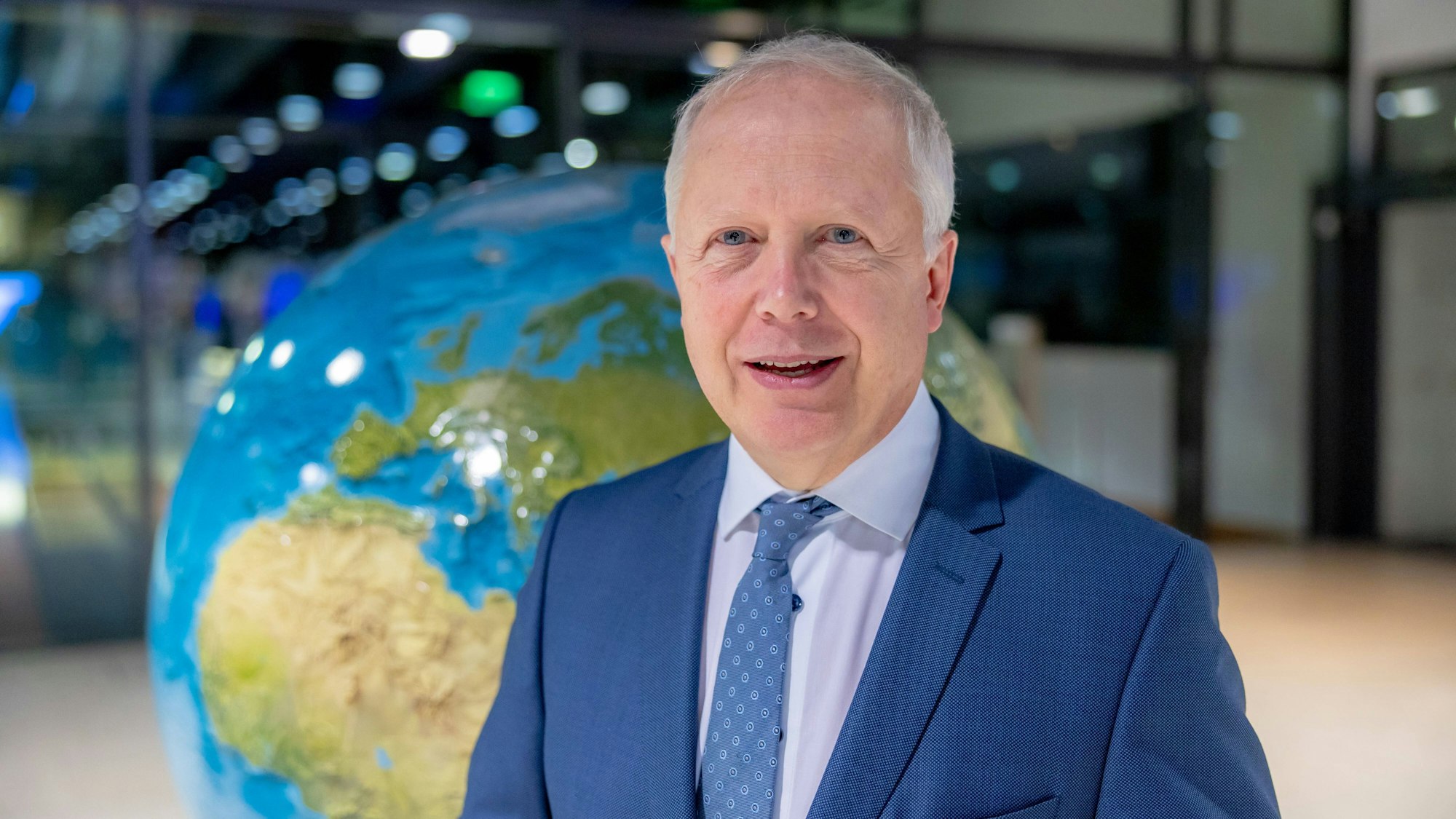 WDR-Intendant Tom Buhrow steht vor einer Weltkugel. Er trägt einen blauen Anzug und lächelt in die Kamera.