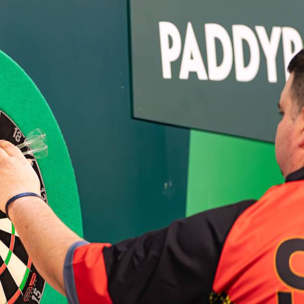Eine Szene aus der laufenden Darts-WM: Der deutsche Spieler Gabriel Clemens sammelt seine Pfeile wieder ein. Er ist im Halbprofil von hinten zu sehen und trägt ein schwarz-orangenes Sportshirt.