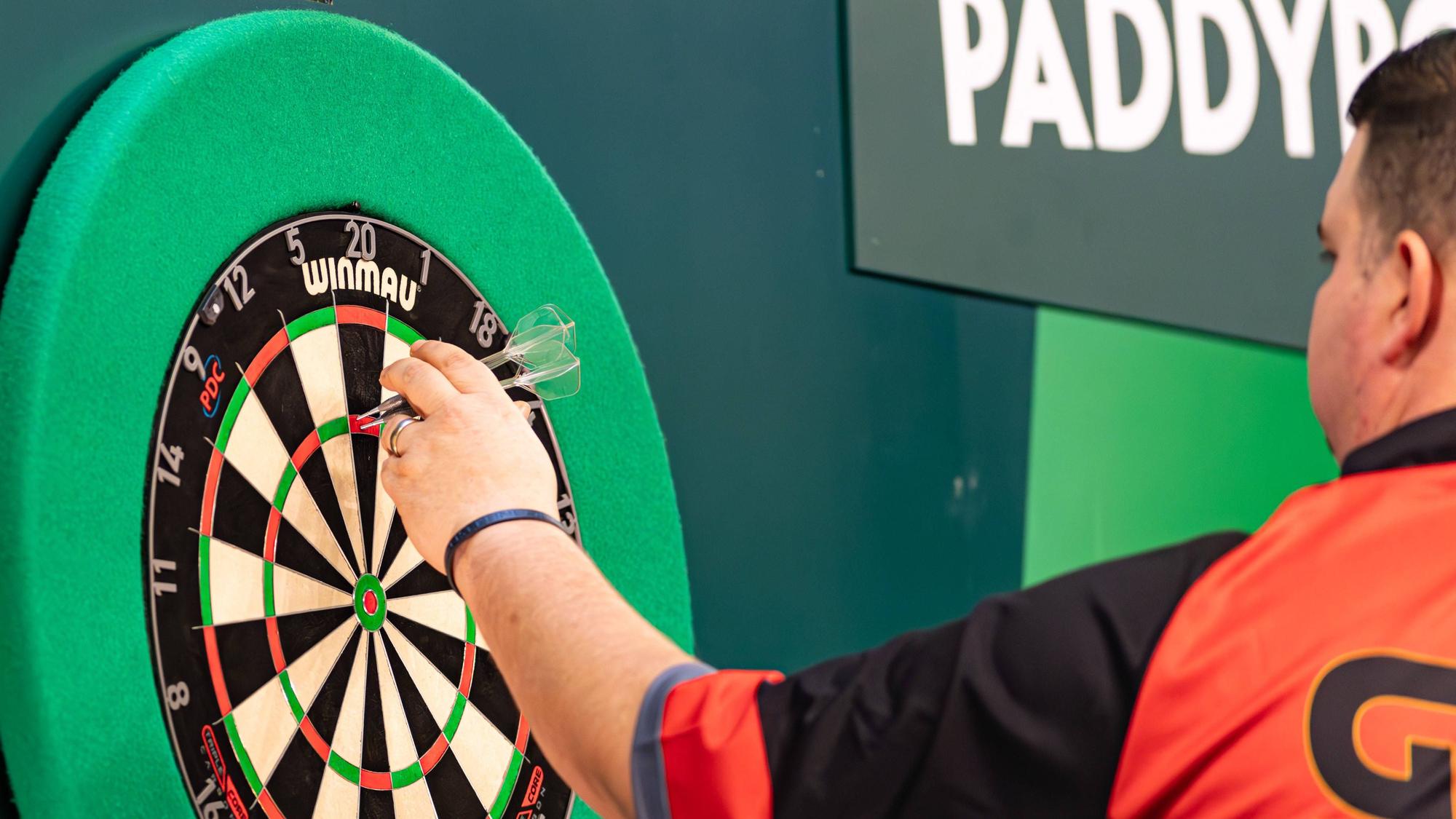 Eine Szene aus der laufenden Darts-WM: Der deutsche Spieler Gabriel Clemens sammelt seine Pfeile wieder ein. Er ist im Halbprofil von hinten zu sehen und trägt ein schwarz-orangenes Sportshirt.