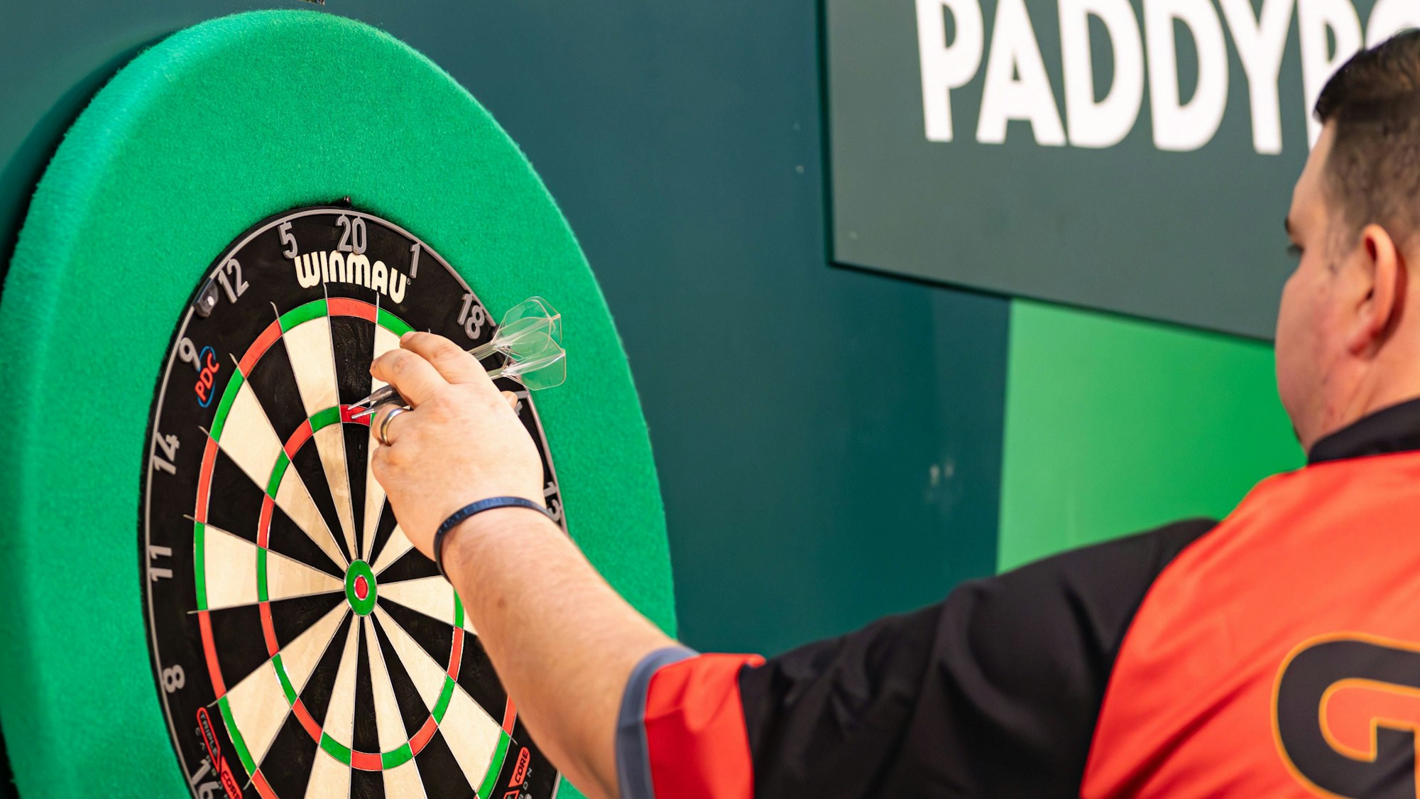 Eine Szene aus der laufenden Darts-WM: Der deutsche Spieler Gabriel Clemens sammelt seine Pfeile wieder ein. Er ist im Halbprofil von hinten zu sehen und trägt ein schwarz-orangenes Sportshirt.