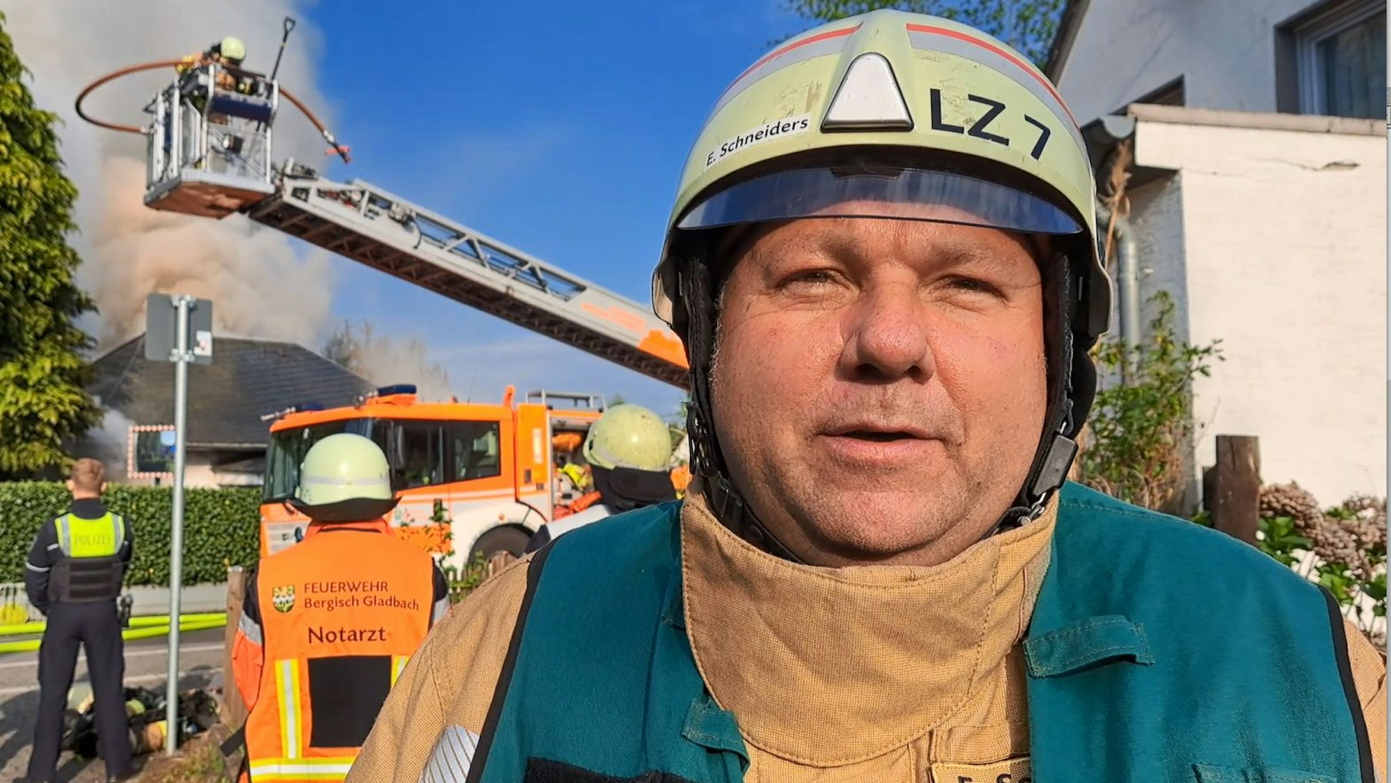 Feuerwehrpressesprecher Elmar Schneiders steht an einer Einsatzstelle, im Hintergrund sind ein brennendes Haus, eine Drehleiter, Feuerwehrleute und ein Polizeibeamter zu sehen.
