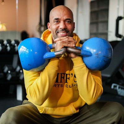 Trainer und Coach Clifford Opoku-Afri sitzt mit zwei blauen Kettlebells auf einer Bank in seinem Fitness-Studio in Ehrenfeld.