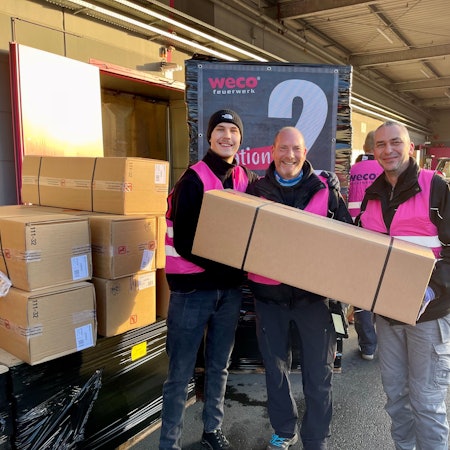 Drei Männer in pinkfarbenen Weco-Westen halten ein langes Überraschungspaket.