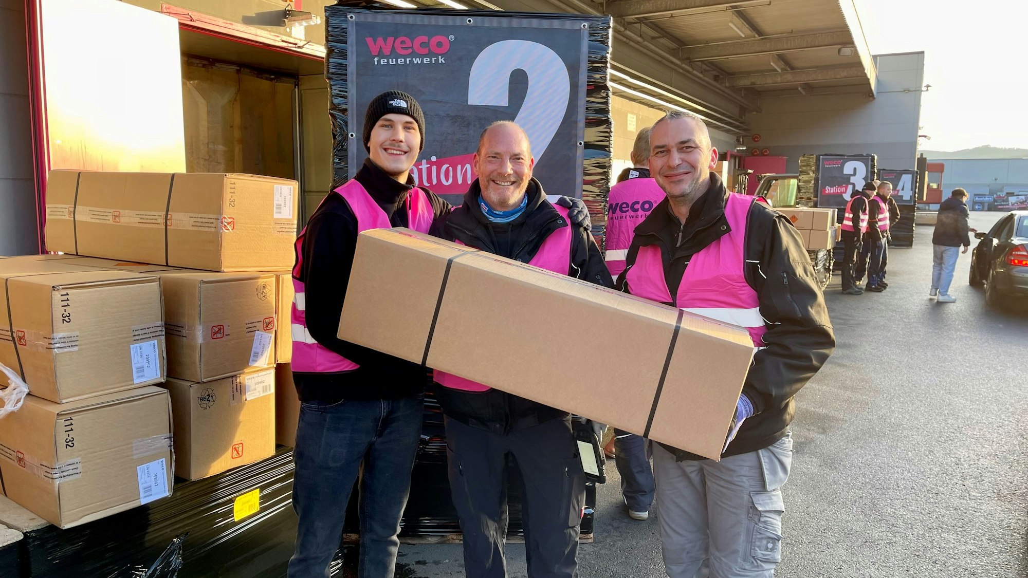 Drei Männer in pinkfarbenen Weco-Westen halten ein langes Überraschungspaket.