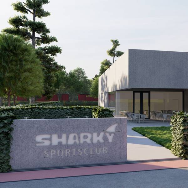 Das Bild zeigt einen modernen Bau, dessen Eingangsbereich mit viel Glas gestaltet ist. Vor dem Gebäude steht ein Stein mit der Aufschrift „Sharky Sportsclub“. An einer Hecke und einer Wiese vorbei führt der Weg zum Eingang.