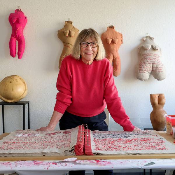 Frau in rotem Pulli in ihrem Atelier vor einem großen, vollgeschriebenen Buch. Die Schrift ist gestickt.