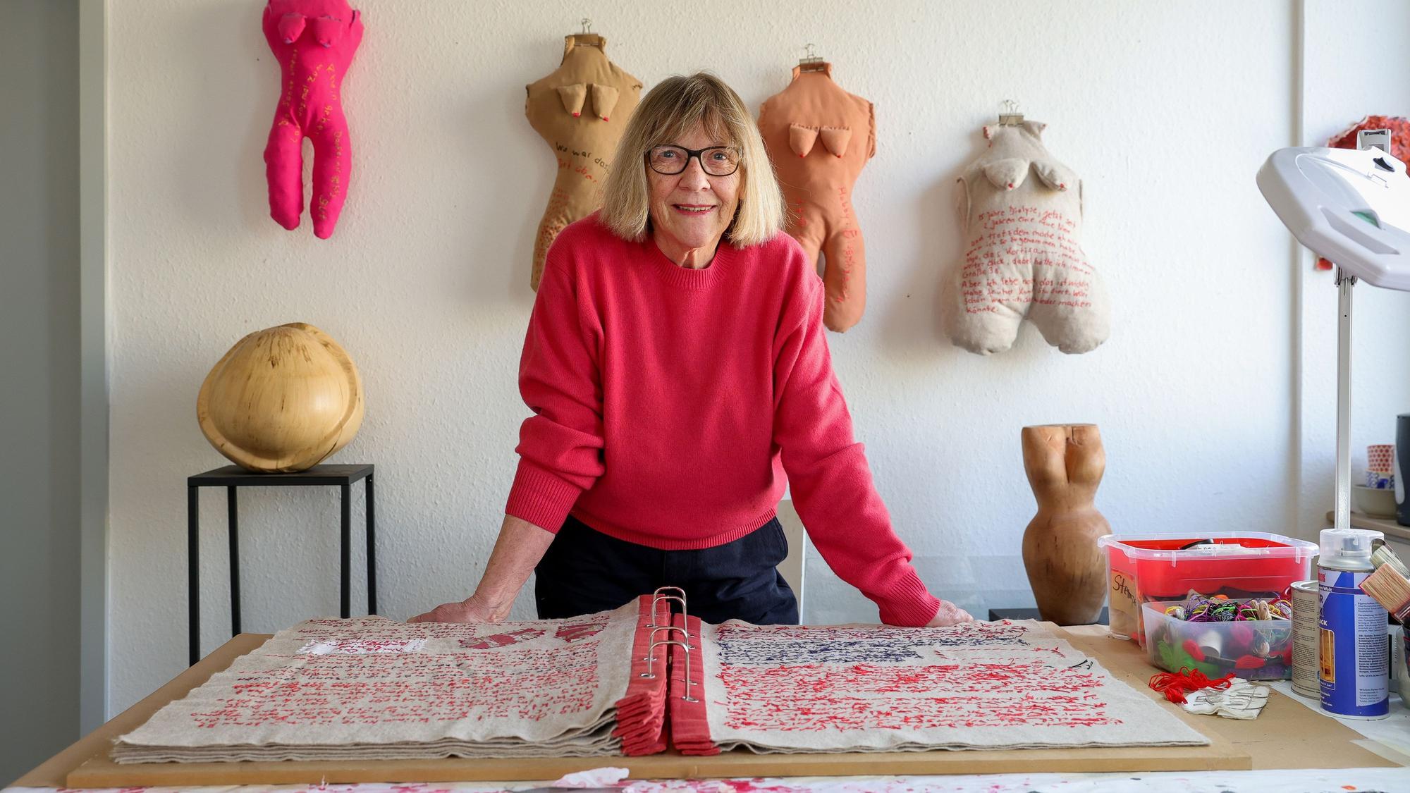 Frau in rotem Pulli in ihrem Atelier vor einem großen, vollgeschriebenen Buch. Die Schrift ist gestickt.