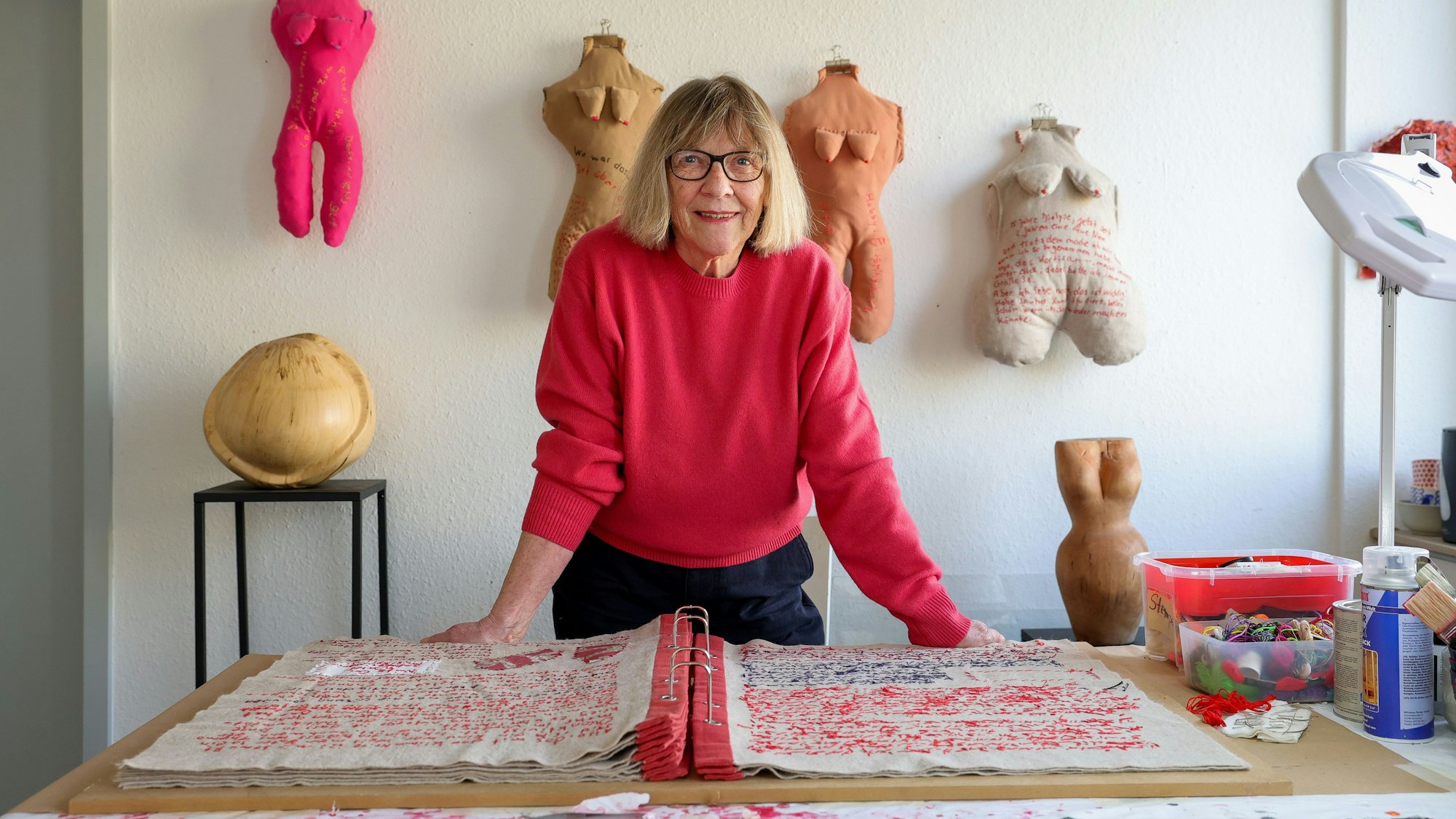 Frau in rotem Pulli in ihrem Atelier vor einem großen, vollgeschriebenen Buch. Die Schrift ist gestickt.